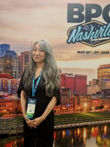 A person with long, wavy hair stands in front of a backdrop featuring a cityscape of Nashville. The backdrop includes the text 'BPC Nashville' and the dates 'May 18th - 21st, 2025.' The person is wearing a black outfit and has a lanyard with an ID badge around their neck