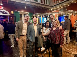Group of five adults posing together inside a lively bar with brick flooring and festive string lights. 