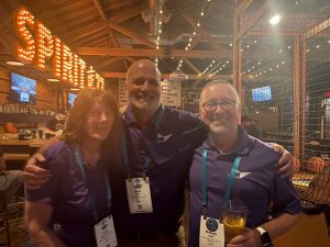 Three colleagues in matching purple shirts smiling with arms around each other at Spirit of 77, string lights and the venue's illuminated sign visible above.