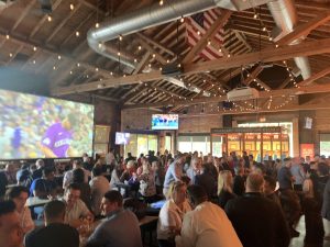 A crowded reception at Spirit of 77, with attendees gathered under string lights near a large screen and TVs mounted on a wood-beamed wall.