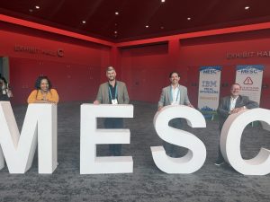 Four colleagues seated at large "MESC" letter sculptures in the exhibit hall, with conference banners visible in the background.