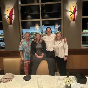 Four people pose shoulder to shoulder facing the camera. They are at a restaurant and behind them is a large window. Kasia is wearing a blue decorative dress, Andrea in a black dress and jacket, Matt in a white shirt, and Tammy in a white shirt and blazer.