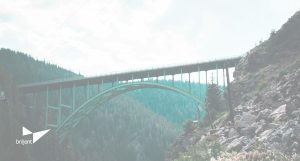 A tall green steel arch bridge spans across a forested mountain valley. Pine trees cover the hillsides, and rocky slopes rise on either side of the bridge. The sky is bright and hazy. In the lower left corner, the Briljent logo appears in white.
