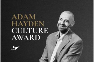 Portrait of a man in a gray suit jacket and checkered shirt beside text announcing the Adam Hayden Culture Award by Briljent.