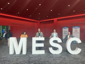 Four colleagues seated at large "MESC" letter sculptures in the exhibit hall, with conference banners visible in the background.